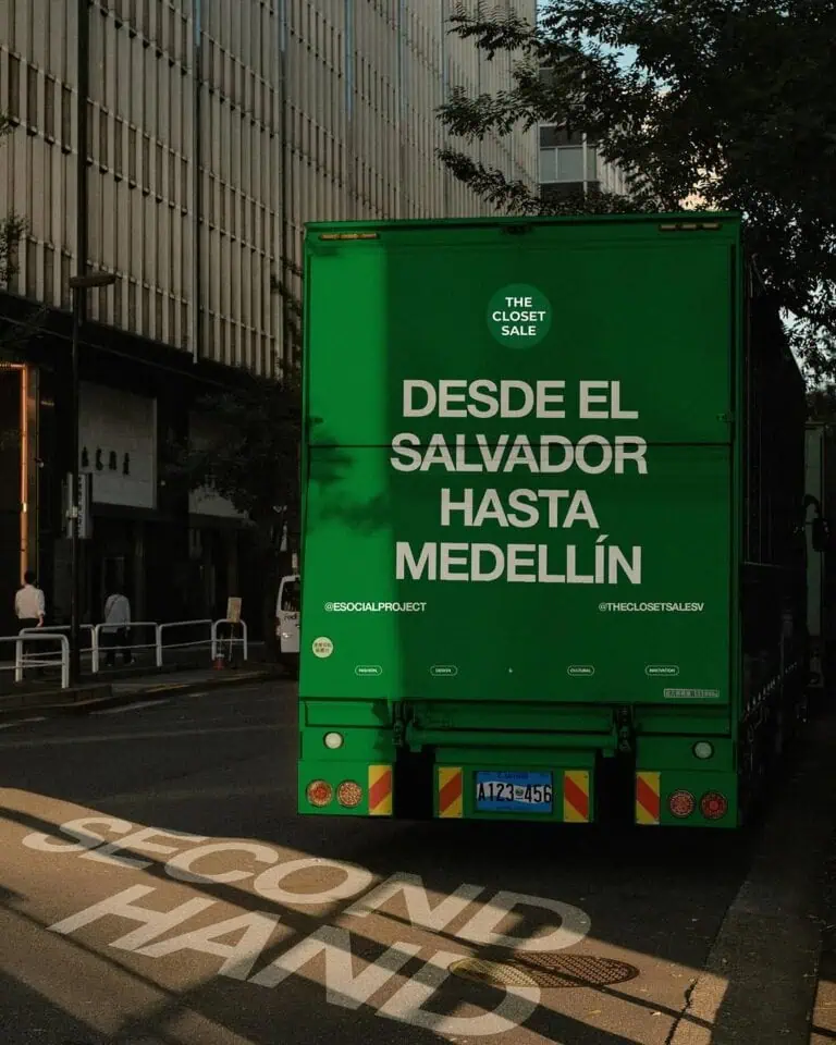 一辆绿色搬家卡车行驶在城市街道上，车身上挂着促销标语，宣传从萨尔瓦多到麦德林的衣物清仓甩卖活动。