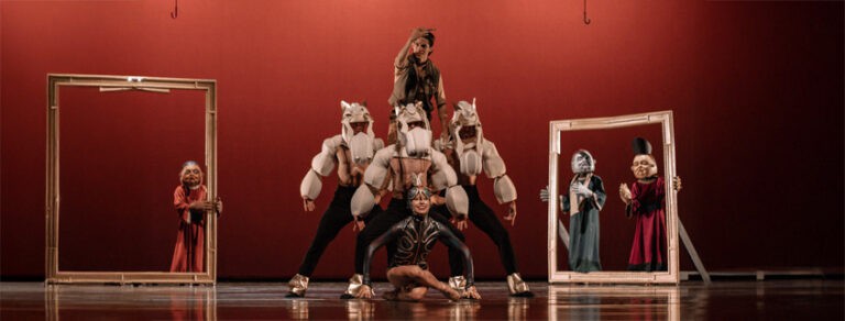Un grupo de artistas en un espectáculo teatral con disfraces y máscaras en un escenario con marcos de madera y fondo color rojo, promoviendo eventos culturales y teatro en Colombia.