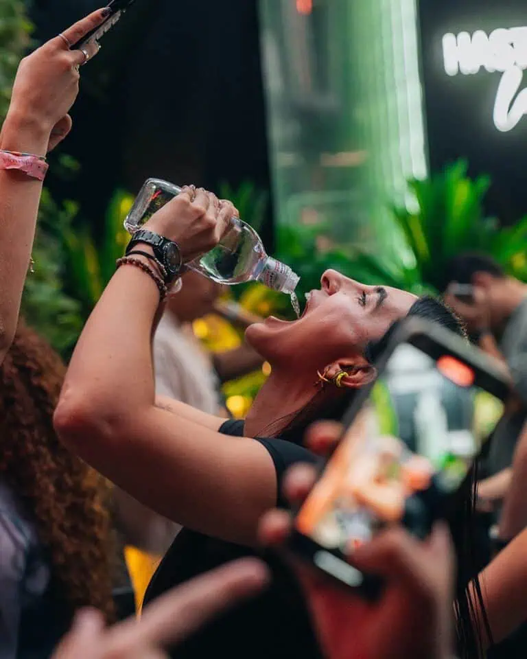 A bottle of water being poured into a woman's mouth at a social event, surrounded by people, in a modern and vibrant environment, promoting hydration and well-being at cultural and business events.