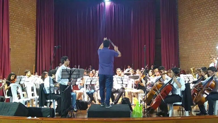 Orchestre symphonique de jeunes en concert dans un théâtre aux rideaux rouges, dirigé par un chef d'orchestre sur scène, des musiciens jouant des instruments à cordes et à vent dans une performance artistique.