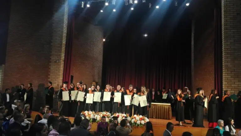 Des diplômés lors d'une cérémonie de remise de diplômes dans un auditorium. Les étudiants, portant toges et casquettes, agitent des drapeaux célébrant leurs réussites académiques. Il s'agit d'un événement institutionnel en Colombie.