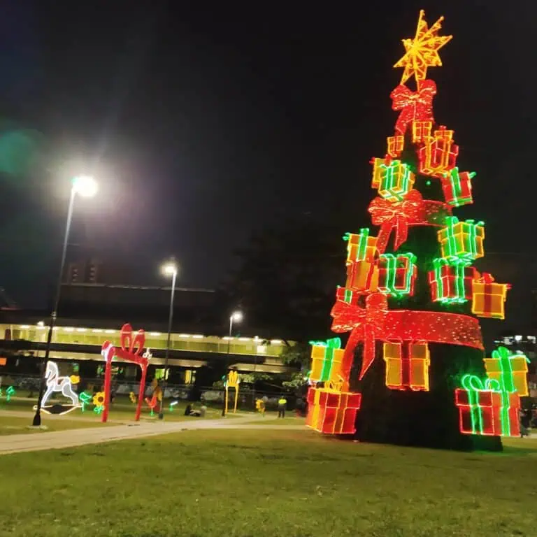 Weihnachtsbaum mit bunten LED-Lichtern und Geschenken in einer städtischen Umgebung bei Nacht.