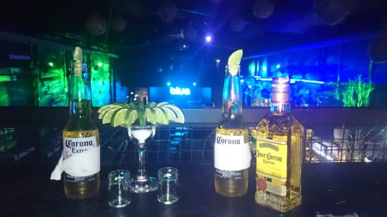 Botellas de cerveza corona y tequila con limón, en un ambiente de fiesta nocturna con luces de colores y música en el fondo.