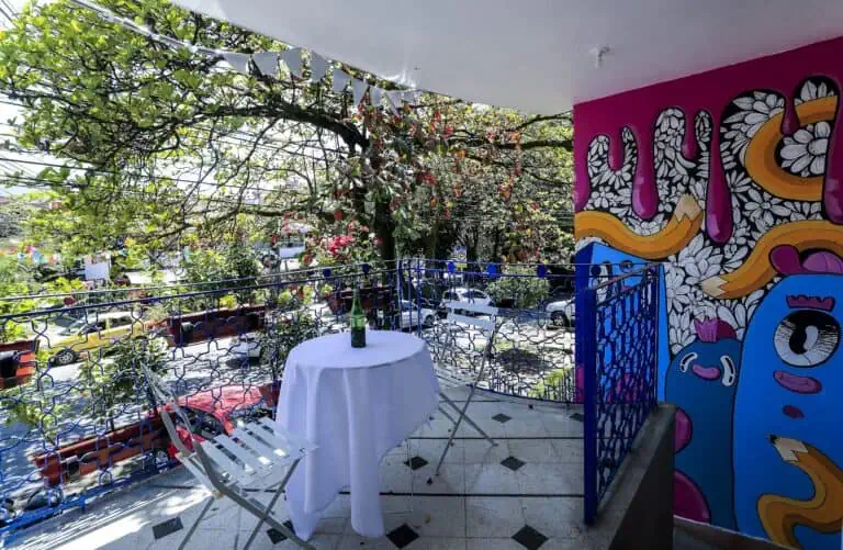 Un balcon décoré d'une fresque murale colorée, une table avec des boissons, des plantes, des arbres et une vue sur la rue lors d'un événement en plein air dans une ville colombienne.