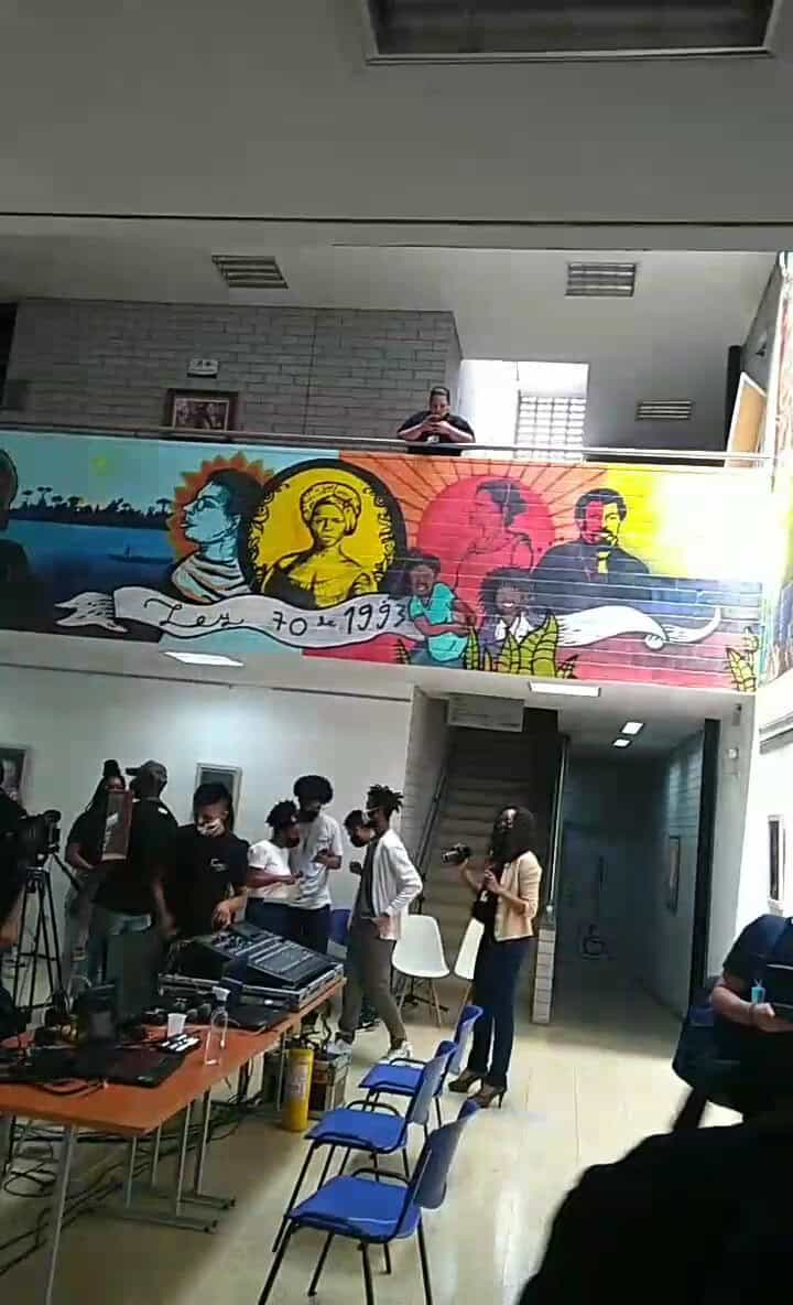 Banda de personas en interior de edificio con mural colorido en la pared, ambiente para evento o reunión, presencia de equipo de sonido y cámaras, espacio de creatividad y cultura, evento social o cultural en Colombia.