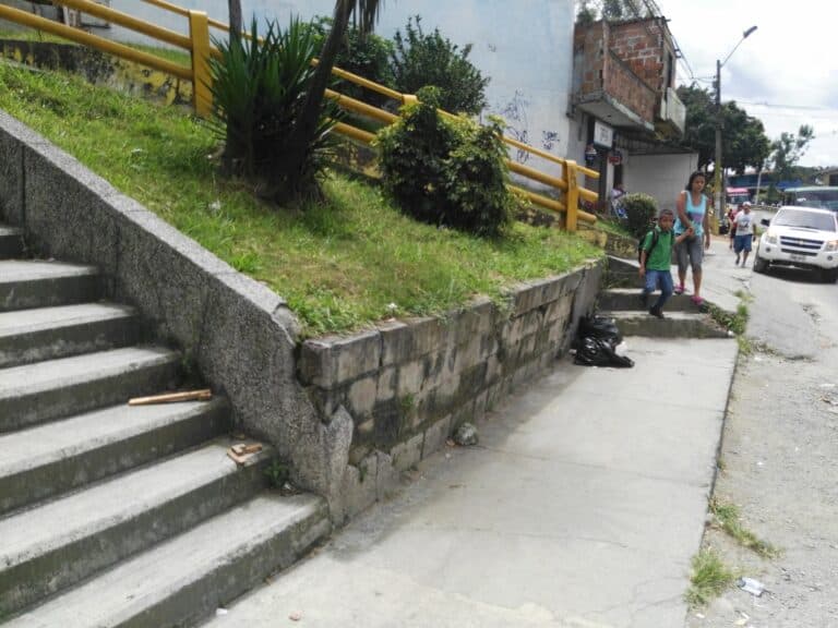 Valla de concreto con escaleras en un barrio urbano. Exactamente en la calle, personas caminando, niños con mochila, y edificios con graffitis y tiendas, reflejando un ambiente cotidiano en un entorno de ciudad.