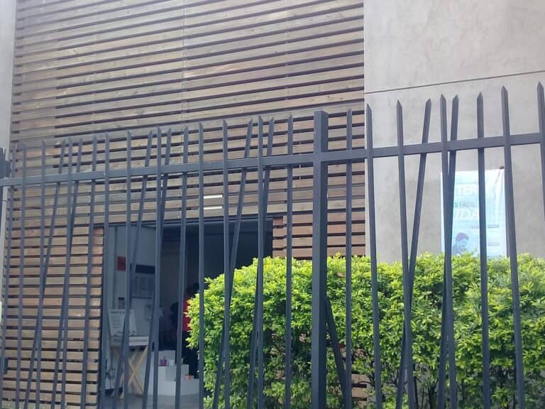 Rejilla de seguridad negra en la entrada de un edificio moderno con fachada de madera y concreto, decorada con arbustos verdes, en un entorno urbano y contemporáneo.