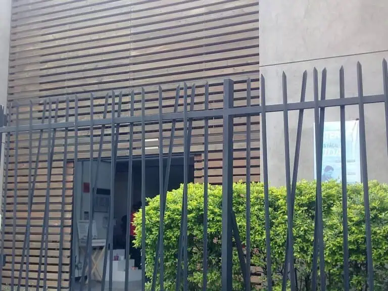 Griglia di sicurezza nera all'ingresso di un edificio moderno con facciata in legno e cemento, decorato con cespugli verdi, in un contesto urbano e contemporaneo.