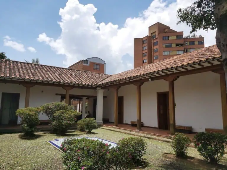 Grande cour intérieure avec jardins et toit en tuiles rouges, entourée d'une structure coloniale avec colonnes en bois et murs blancs, dans un cadre urbain avec des bâtiments modernes en arrière-plan.