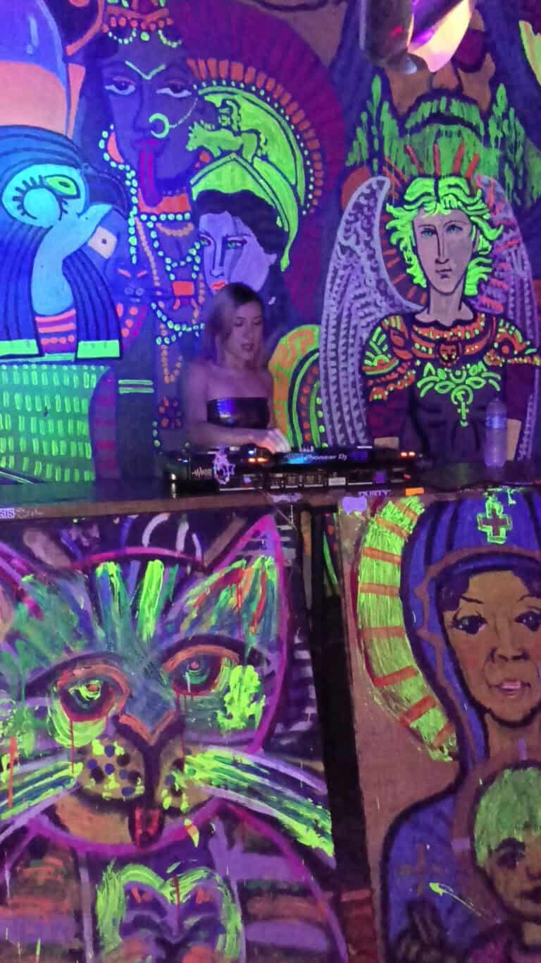 Mujer DJ en un evento con arte callejero fluorescente y pinturas neón que resaltan en la oscuridad, representando un ambiente artístico y vibrante en la celebración.