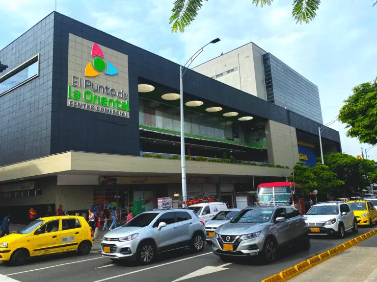 Punto di incontro nel centro commerciale La Oriental in una giornata di sole, con traffico automobilistico e pedonale, a dimostrazione della modernità e dell'attività commerciale in un ambiente urbano in Colombia.