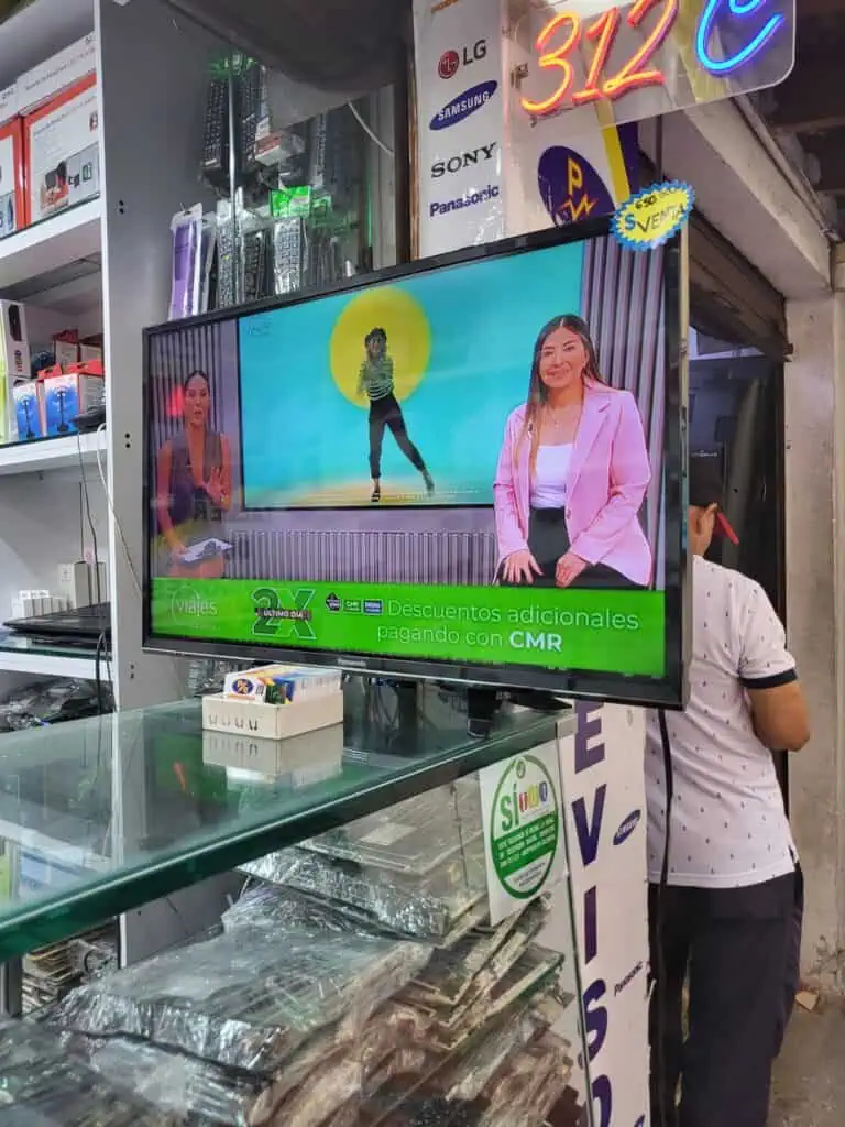 在哥伦比亚一家商业环境中，一家电子商店的电视屏幕上播放着旅游和科技产品的折扣信息，背景色彩鲜艳，文字为西班牙语。