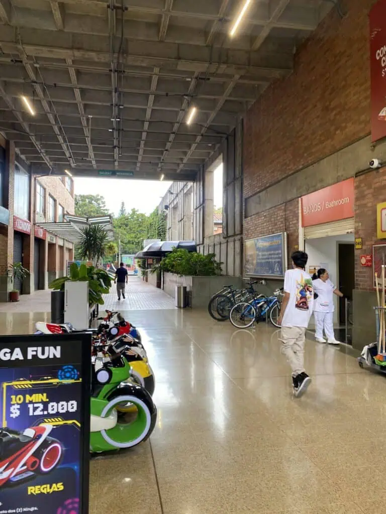 Alttext: Innenkorridor eines Einkaufszentrums mit Elektrorollern, Toiletten- und Fahrradbeschilderung, moderne und helle Atmosphäre, Servicebereiche in Kolumbien.