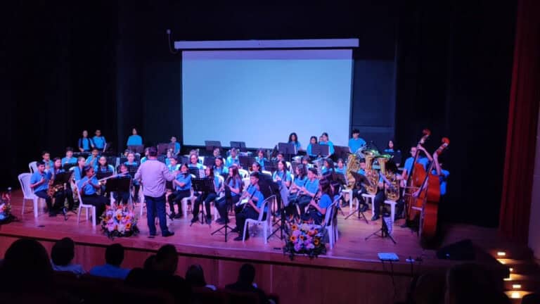 Joven orquesta de músicos en concierto en un escenario teatral, dirigida por un maestro, con instrumentos como saxofones, clarinetes, y contrabajo, en un evento cultural destacado.
