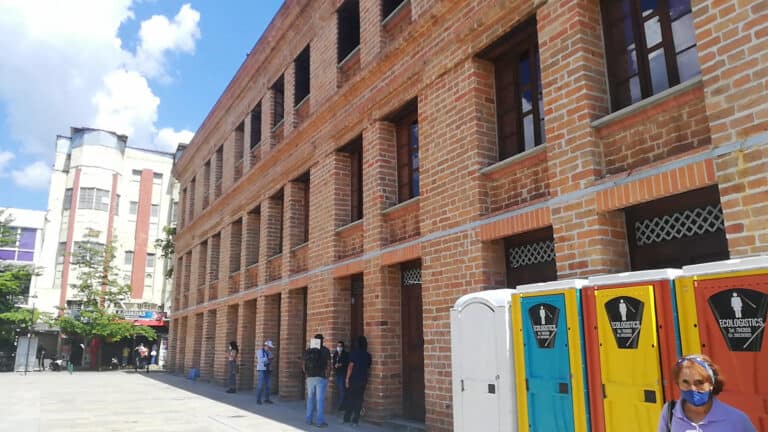 Edificio de ladrillos rojos en un entorno urbano con personas usando mascarillas y contenedores de reciclaje en primer plano, representando un escenario de eventos o infraestructura en Colombia.
