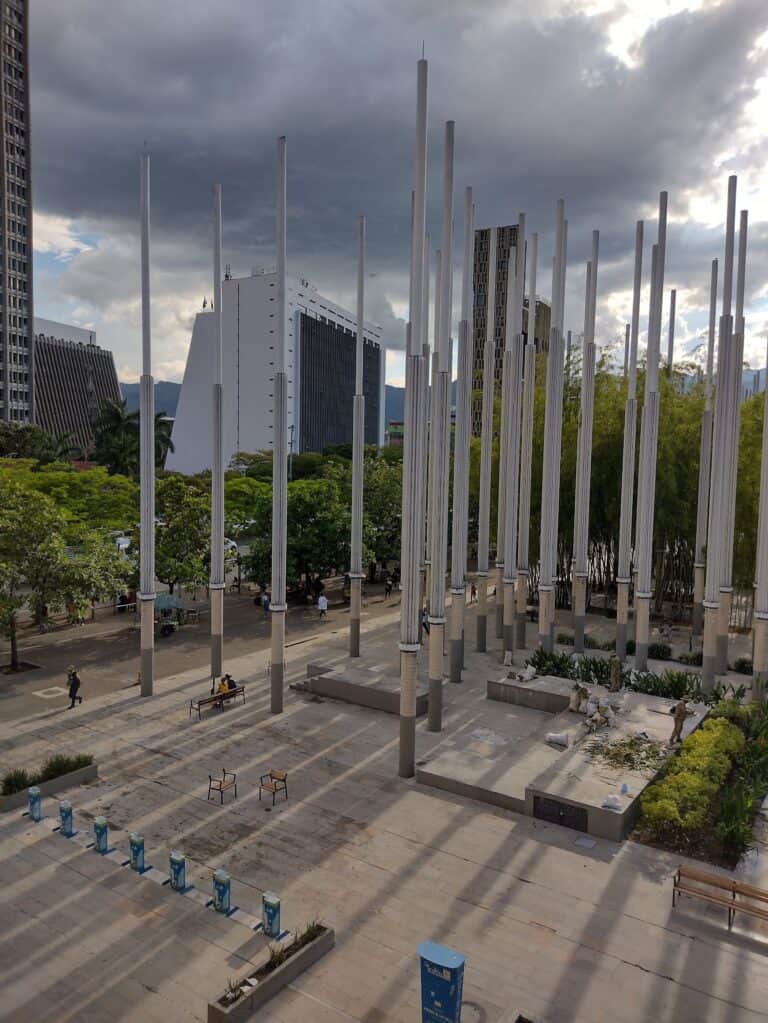 Jardín urbano con columnas metálicas en una plaza, rodeado de edificios modernos y árboles verdes, con cielo nublado, ideal para eventos corporativos y festivales en la ciudad de Bogotá.