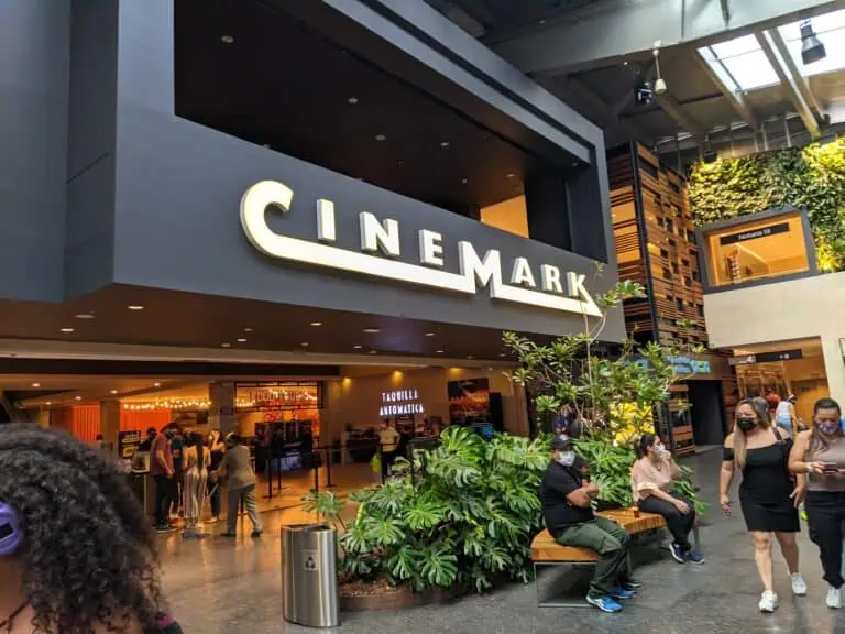 Abweichung: Eingang zu einem Kino in einem Einkaufszentrum mit maskierten Menschen und dekorativen Pflanzen darin.