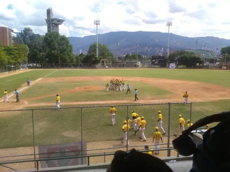 Baseball players in a game on an outdoor sports field, with the team wearing yellow uniforms, in an urban setting with mountains in the background and a sports lighting structure.