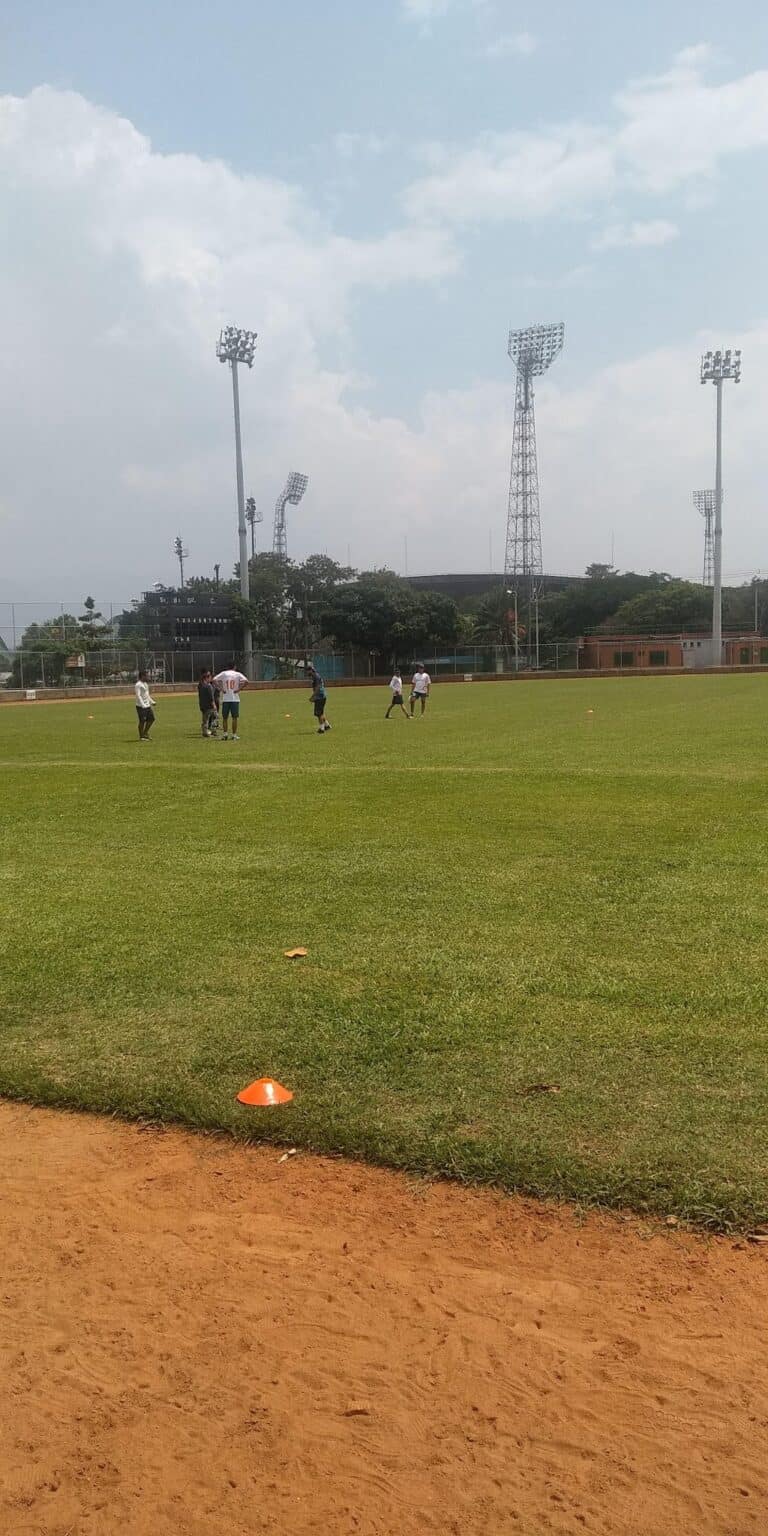 Entrenamiento de fútbol en campo abierto con jugadores practicando en un césped amplio y bien cuidado, con torres de iluminación y estadio en el fondo, bajo cielo nublado.