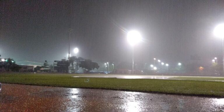 Campo de fútbol iluminado con torres de luz en la noche lluviosa, reflejándose en el césped y el suelo mojado. Escenario ideal para eventos deportivos y actividades al aire libre.