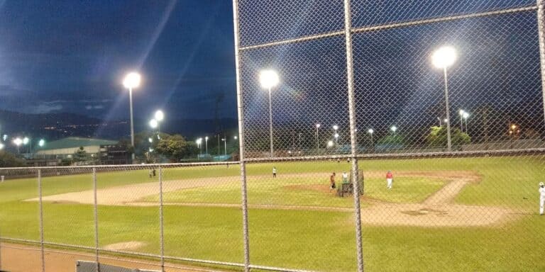 Campos de béisbol iluminados en la noche, rodeados con valla metálica y con jugadores en el campo.