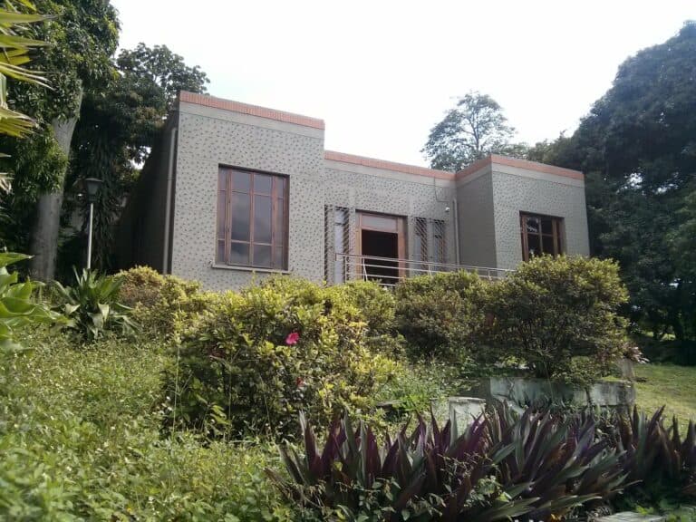 Casa moderna en medio de jardines verdes y árboles, con amplias ventanas de vidrio y un balcón, ideal para eventos al aire libre y reuniones en un entorno natural.