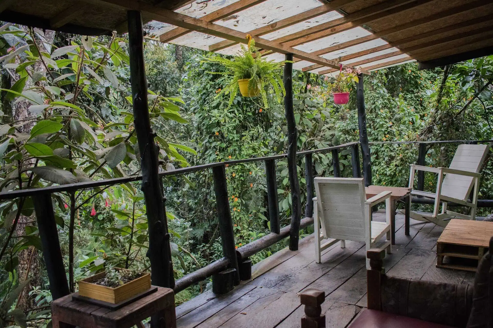 Ein Balkon inmitten der Natur, mit weißen Stühlen und rosa und gelben Hängepflanzen, umgeben von üppiger Vegetation in einer ruhigen, natürlichen Umgebung.