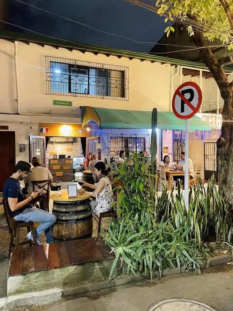 夜晚的城市露天咖啡馆和餐厅，人们在绿树环绕、禁止停车的标志下享用饮品和食物。