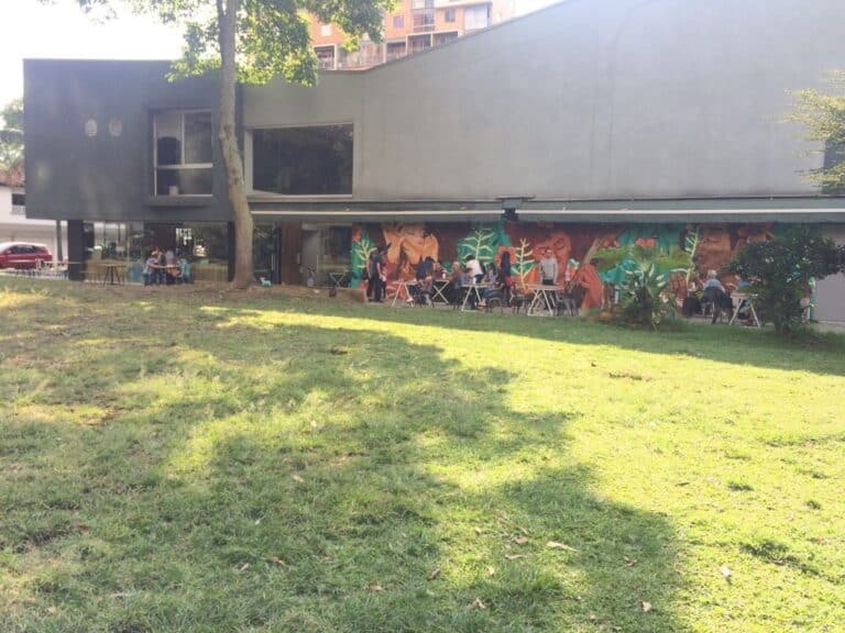 Modern building with outdoor seating area on green lawn, bustling with people enjoying social events and dining, featuring vibrant mural artwork on exterior wall, surrounded by trees and urban environment.