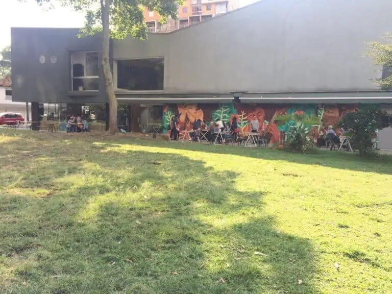 现代建筑，绿茵茵的草坪上设有户外座位区，人们在此享受社交活动和用餐，外墙上绘有充满活力的壁画，周围环绕着树木和都市环境。