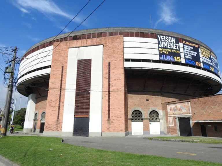 Edificio con cartelera digital de eventos, teatro o conciertos en un espacio público, fachada moderna de ladrillos y estructura circular, en un día soleado con cielo azul claro.