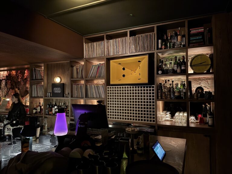 Bar de estilo moderno con estanterías de madera llenas de libros, botellas de alcohol y objetos decorativos en un ambiente con iluminación cálida y ambiente de evento social.