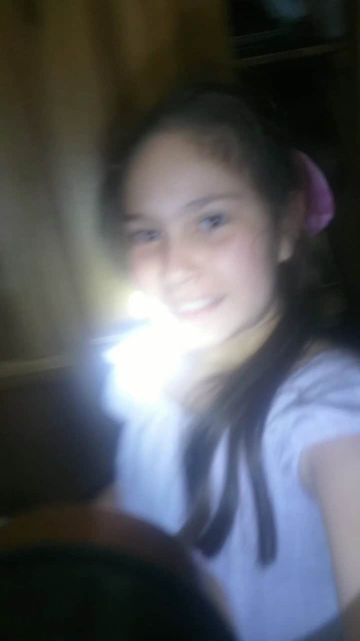 Sonriente niña con sonrisa natural, cabello largo y accesorios rosados, en un ambiente interior con luz natural y fondo de madera, en una foto casual y espontánea.