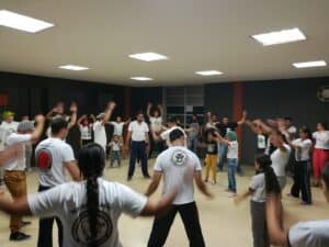 Capoeira infantil en actividad grupal en espacio cerrado, demostrando movimiento y coordinación en taller para niños y adultos en evento comunitario.