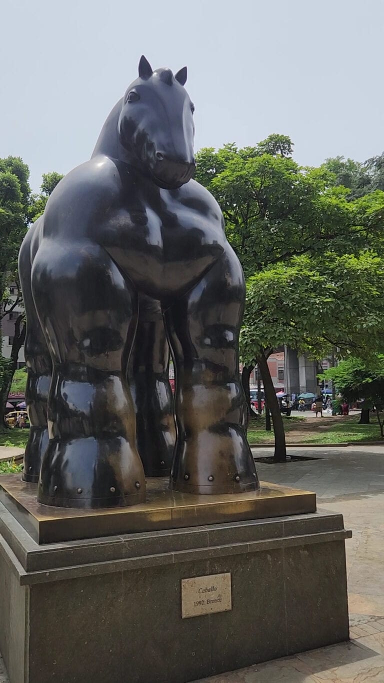 Bufalo de bronce, escultura moderna ubicada en un parque con árboles y naturaleza en el fondo, obra artística de 1992, simboliza fuerza y dominio en espacios públicos.