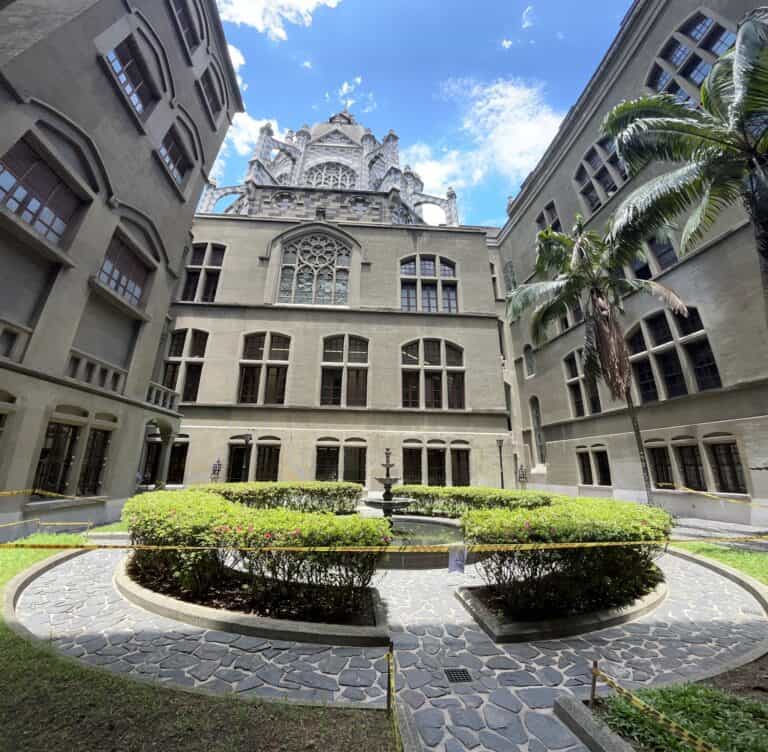 Imponente edificio histórico en medio de un patio ajardinado con fuente, rodeado de palmeras y ventanas grandes, bajo un cielo azul con algunas nubes, ideal para eventos y reuniones culturales.
