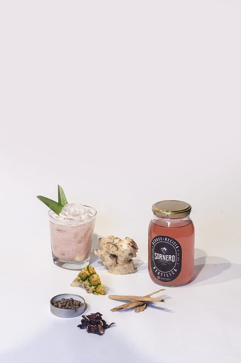 Innovador cóctel saludable en un vaso con hielo y una rodaja de piña, acompañado de ingredientes naturales como jengibre, pimienta negra, y hierbas, promoviendo un estilo de vida sostenible y eco-amigable en eventos.
