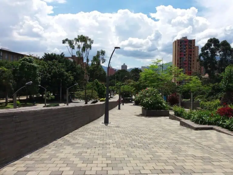 Een breed wandelpad in een stadspark met bomen en sierplanten, ideaal voor evenementen en activiteiten in de stad, met woongebouwen en een bewolkte lucht op de achtergrond.
