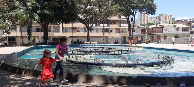 在城市公园的喷泉旁，婴儿们奔跑嬉戏，背景是树木和建筑物，呈现出一幅热闹而温馨的城市景象。