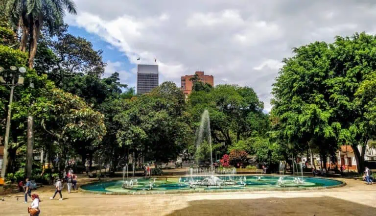 Ein Garten mit einem Brunnen und Bäumen im Stadtzentrum mit hohen Gebäuden im Hintergrund, ein belebter grüner öffentlicher Raum an einem sonnigen, bewölkten Tag.