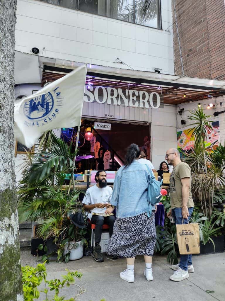 Flamante bandera del club social en la entrada del bar SORNERO, con personas conversando y disfrutando del ambiente en la vía pública, rodeados de plantas y decoración moderna.