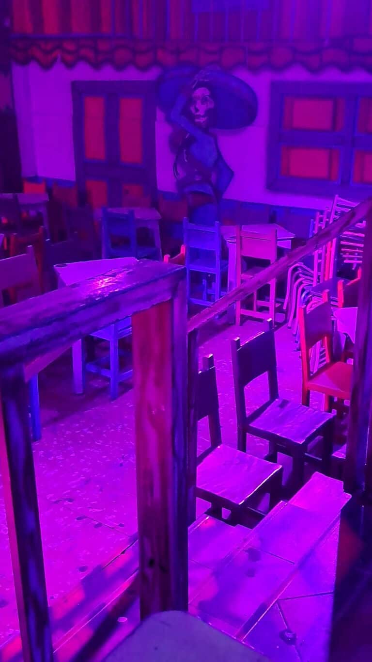 Mesas y sillas en un restaurante con iluminación de luces violetas y decoraciones tradicionales mexicanas.