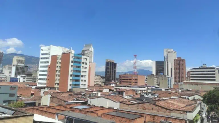 Eine weite Stadtansicht von Medellín mit modernen und traditionellen Gebäuden unter klarem Himmel, die den Blick auf die Stadt und ihre Berge im Hintergrund widerspiegelt.