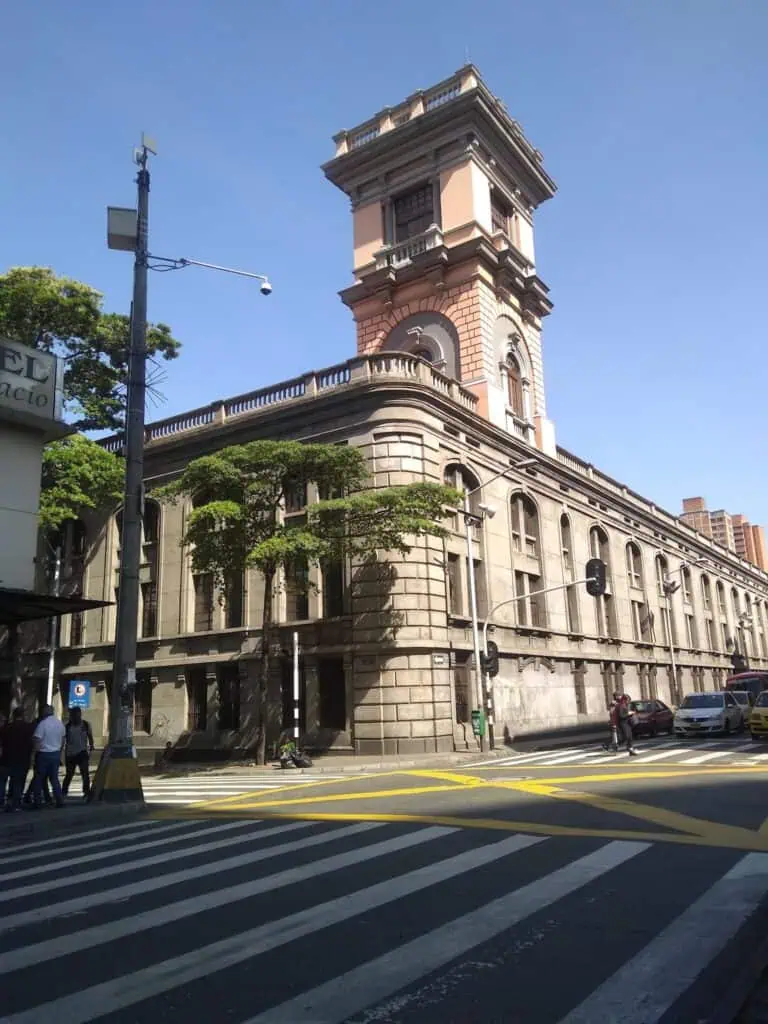 波哥大市中心一座古老的古典风格建筑，带有塔楼，建筑细节以粉色和灰色为主，周围环绕着树木和交通标志，天空晴朗。