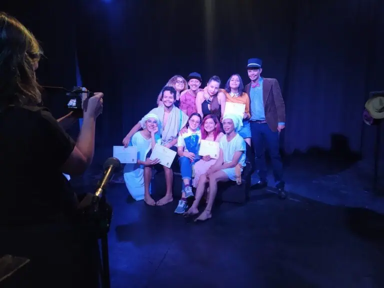 Des étudiants et des acteurs présentent des certificats lors d'un événement théâtral, célébrant les réalisations académiques et artistiques en costumes de théâtre, sur une scène sombre avec des projecteurs.