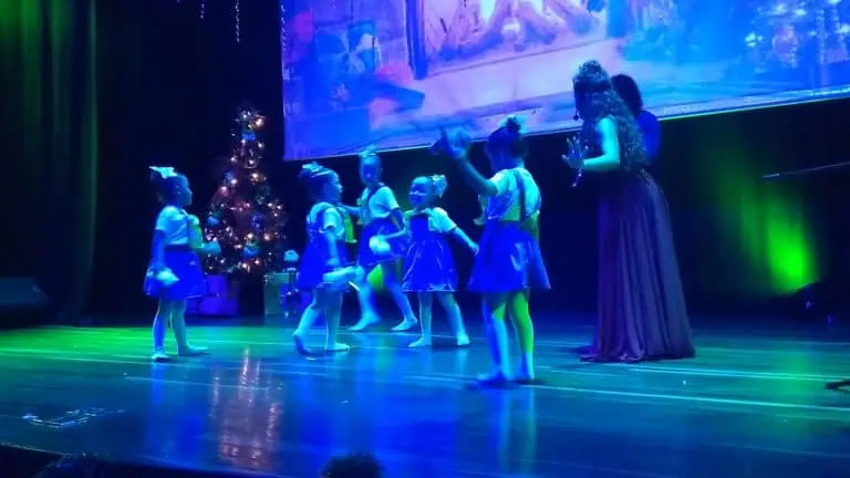 Ragazze che si esibiscono su un palco illuminato con luci colorate e decorazioni natalizie, in occasione di un evento per bambini o scolastico organizzato da Eventario.