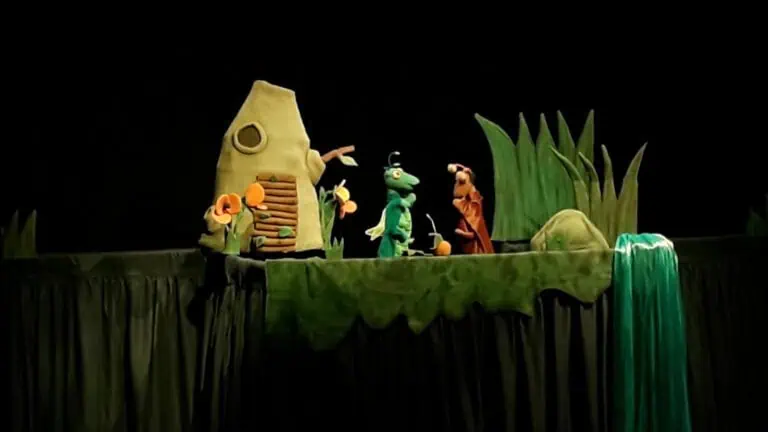 Eine Marionettenpuppe mit Figuren aus einem Kindertheaterstück auf der Bühne, mit einer Naturkulisse mit Haus, Pflanzen und Wasserfall, ideal für Veranstaltungen und Kindertheater.