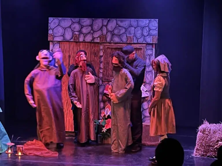 Theaterkostüme von Figuren aus Kinderbüchern in einer Live-Aufführung mit einer dekorierten Bühne, die ein Haus aus Stein und Holz simuliert, mit Schauspielern in farbenfrohen Kostümen und lustigen Gesichtsausdrücken.