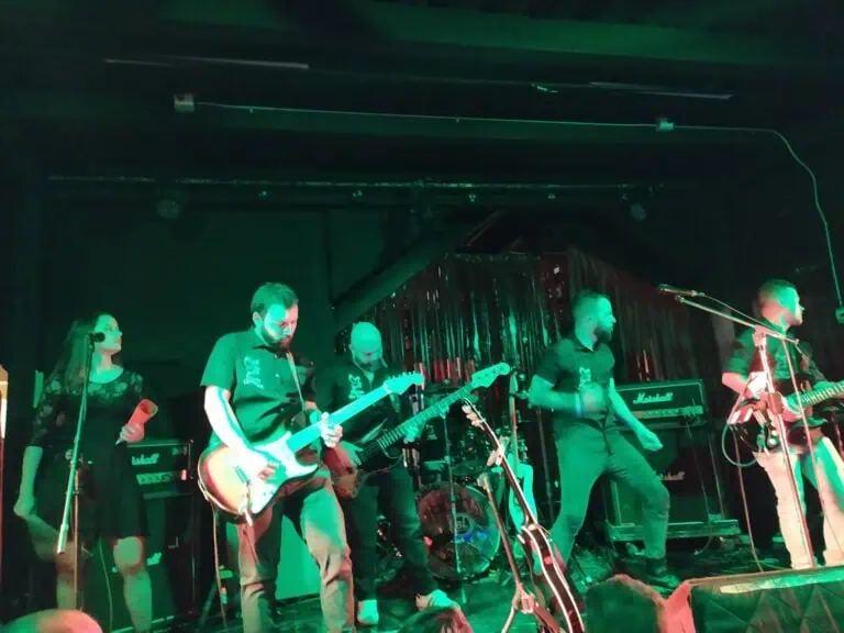 Live-Musikband bei einem Konzert auf einer Bühne mit grüner Beleuchtung und schwarzem Vorhang im Hintergrund.