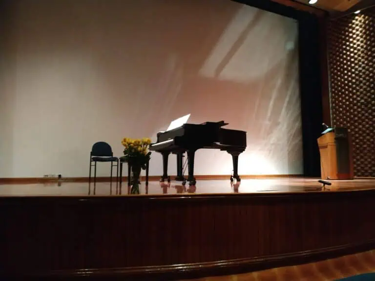 Ein Bühnenklavier für kulturelle Veranstaltungen und Online-Konzerte mit Ambientebeleuchtung, dekoriert mit einer Stuhllehne, einer blühenden Pflanze und einem Podium, bereit für Präsentationen und künstlerische Aktivitäten in Kolumbien.